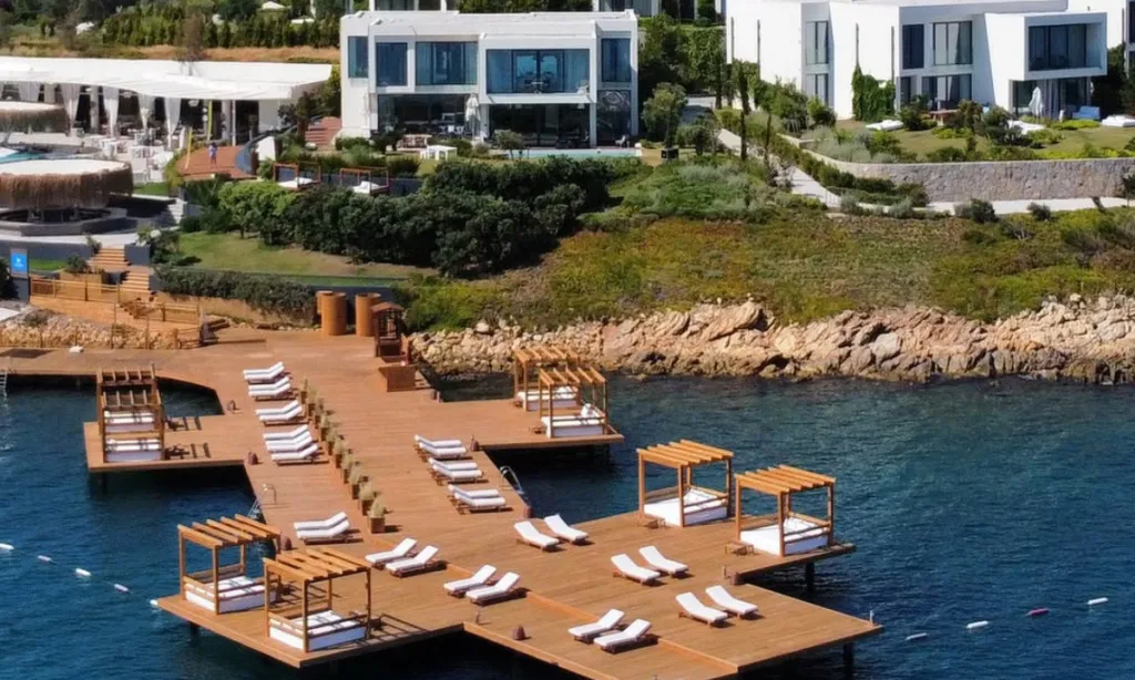Bodrum seyahat rehberi: Suyah Pool Club'ın havadan çekilmiş bu geniş görüntüsünde, masmavi denizin üzerine uzanan uzun ahşap iskelede sıralanmış beyaz şezlonglar, gölgelikli daybed locaları ve arka planda yemyeşil yamaçlara serpiştirilmiş lüks modern binalar görünüyor.