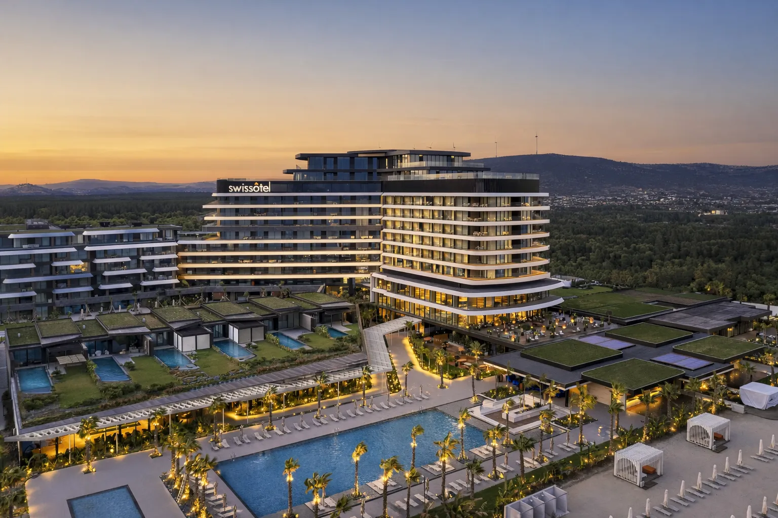 Gün batımında çekilmiş bir hava fotoğrafında, ışıkları yanan modern mimarili Swissôtel Resort & Spa Çeşme oteli, yemyeşil bahçeleri, berrak havuzları, palmiye ağaçları ve geniş dinlenme alanlarıyla göz alıcı bir şekilde uzanıyor.