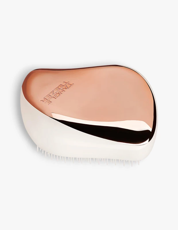 Parlak gül altın rengi üst kısmı ve alt kısmında görünen beyaz esnek dişleriyle, saçları nazikçe dolaşıklıklardan arındıran ergonomik tasarımlı Tangle Teezer Detangling Compact Styler saç fırçası, sade beyaz bir arka plan üzerinde dikkat çekiyor.