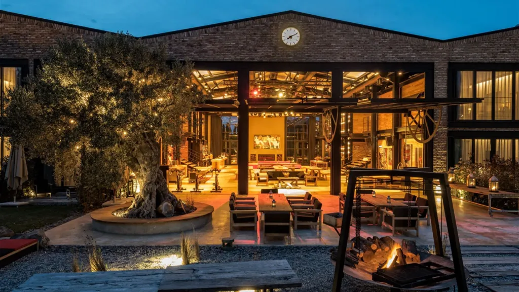 The Stay Warehouse'ın tuğla ve geniş cam cepheli binası, dışarıdan bakıldığında içerideki hareketli yaşam alanlarını gösterirken, avlusundaki peri ışıklarıyla aydınlatılmış yaşlı zeytin ağacı, dış mekan oturma düzenleri ve yanan ateş çukuruyla misafirlerine hem endüstriyel estetiği hem de sıcak bir konaklama deneyimi sunuyor.