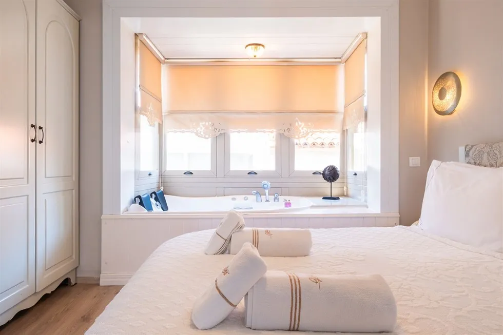 Aydınlık ve modern bir otel odasında, beyaz nevresimli yatağın üzerinde kahverengi çizgili dört rulo havlu dururken, yatağın solunda beyaz bir gardırop ve sağında ışıklı bir duvar apliği görülüyor; odanın arka kısmında, üç pencereli nişte entegre jakuzi ve şık dekoratif objeler ferah bir konaklama deneyimi sunuyor.