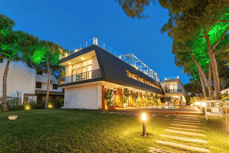 Çeşme'de bulunan Villa Kore'nin modern ve şık mimarisi, çam ağaçlarının yeşil ışıklarla aydınlattığı geniş bahçesinde, üzerinde yürüyenleri karşılayan taş patika lambaları ve su kenarındaki açık oturma alanıyla akşamın mavi gökyüzü altında parlıyor.