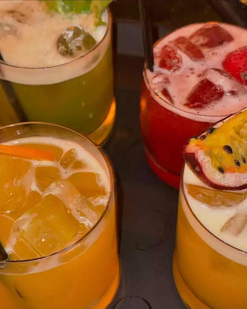 Dört farklı renkteki soğuk içecek bardağı koyu renk bir zeminde yan yana duruyor; içeceklerden biri yeşil nane veya misket limonu dilimli, diğeri çilekli kırmızı, bir başkası portakal dilimli turuncu ve sonuncusu ise yarım çarkıfelek meyvesi ile süslenmiş açık sarı renkte olup hepsinde bolca buz ve köpük bulunuyor.