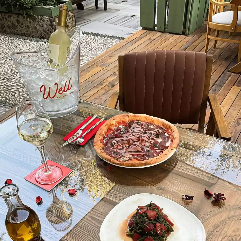 Açık hava bir restoran terasında, Well markalı buz kovasında serinletilmiş beyaz şarap, kadehte beyaz şarap, bol pastırma ve koyu sos gezdirilmiş büyük bir pizzanın, yanında yeşil sebzeler ve kırmızı domates dilimleri içeren bir garnitürün ve zeytinyağı şişesinin bulunduğu şeffaf cam bir masa gösteriliyor.