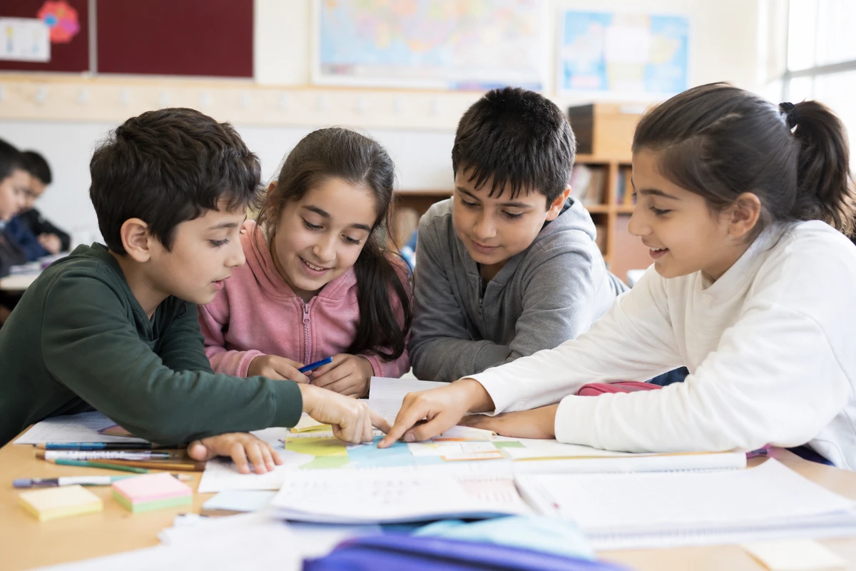 Sınıfta bir masanın etrafında toplanmış, gülümseyen dört ilkokul öğrencisi, renkli bir çalışma üzerinde parmaklarıyla işaretleşerek neşeyle iş birliği yaparken, bu tür sınıf içi iş birliği faaliyetlerinin akran zorbalığının önlenmesinde pozitif etkileşimleri teşvik ettiğini gösteriyor.