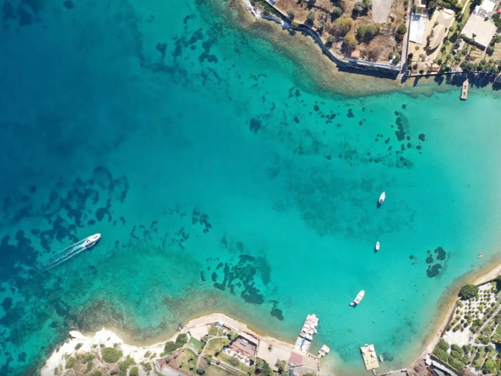 Çeşme'nin Aya Yorgi Koyu'nun havadan çekilmiş bu nefes kesici görüntüsünde, cam gibi berrak turkuaz sular, su altı yaşamının zenginliğini gösteren kayalık ve kumlu alanlarla iç içe geçerek uzanıyor; bir sürat teknesi arkasında köpüklü bir iz bırakırken, sahile yakın demirlemiş tekneler ve yeşil kıyı şeridi Ege'nin dingin güzelliğini vurguluyor.
