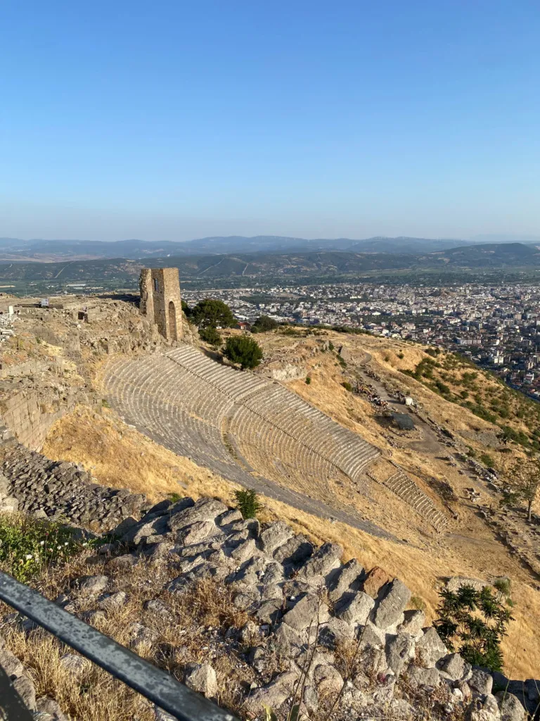 Yüksek bir noktadan çekilen bu panoramik fotoğrafta, kurumuş otlarla kaplı yamaca oyulmuş, kademeli oturma sıralarına sahip görkemli Bergama Antik Tiyatrosu, sağ tarafında İzmir'in Bergama ilçesinin modern yerleşimini ve uzaktaki tepeleri kapsayan geniş bir manzara ile Türkiye'deki bir UNESCO Dünya Mirası alanı olan Bergama Çok Katmanlı Kültürel Peyzaj Alanı'nın zenginliğini gözler önüne seriyor.