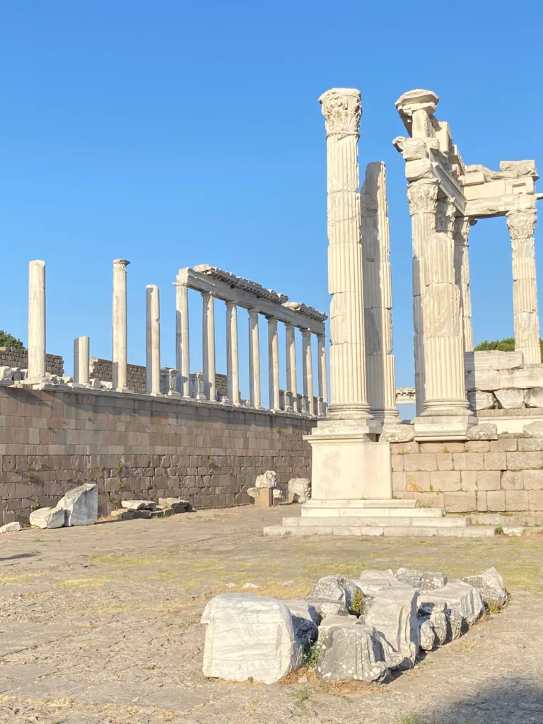 Açık mavi bir gökyüzünün altında, Türkiye'nin zengin kültürel mirası içinde yer alan Bergama Akropolü'ndeki Trajan Tapınağı'na ait, kısmen ayakta duran ve etrafa saçılmış zarif mermer sütunlar ile antik yapı kalıntıları, bu UNESCO Dünya Mirası alanının görkemli geçmişini etkileyici bir şekilde sergiliyor.
