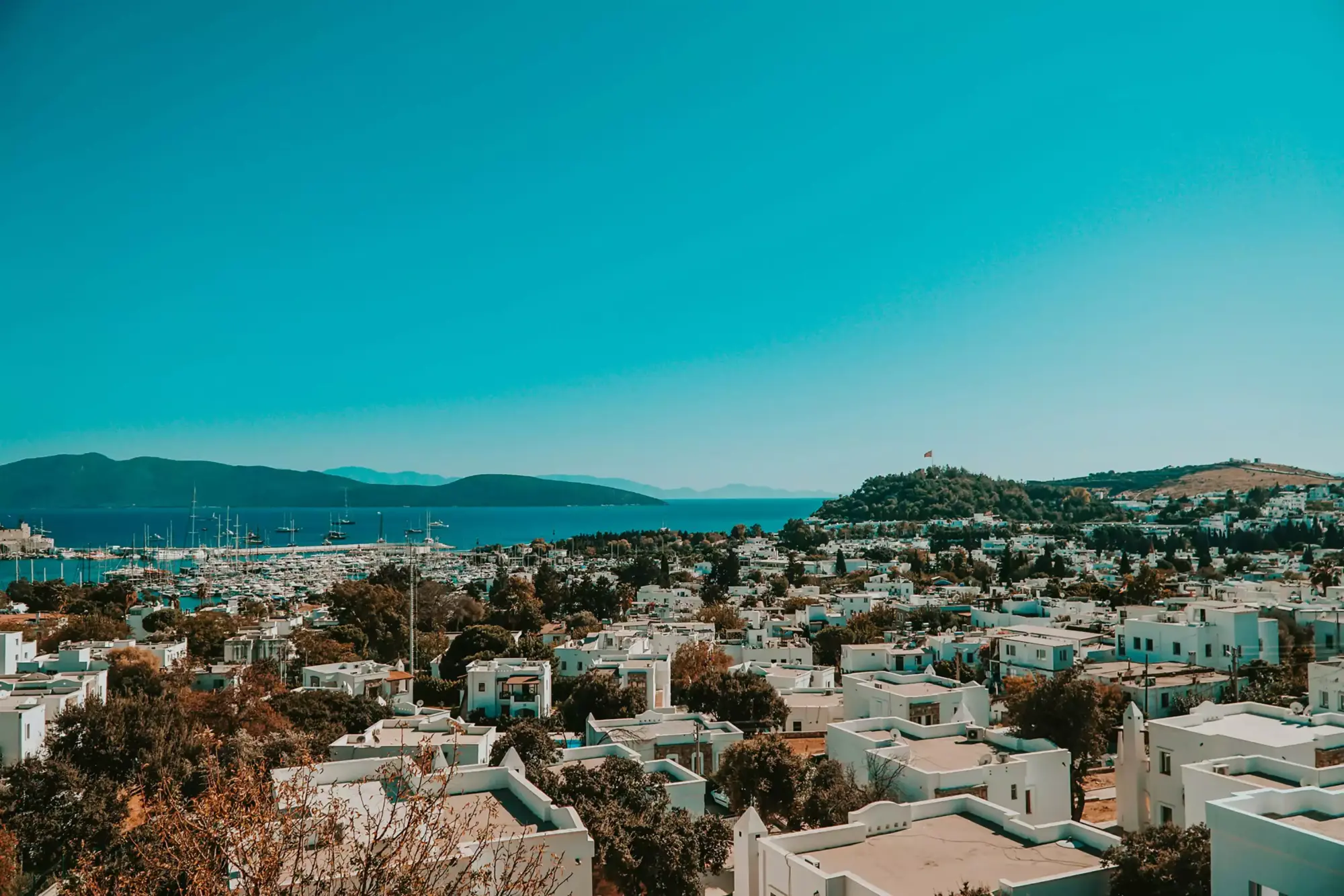Bodrum seyahat rehberi: Bodrum'un ikonik manzarası; masmavi berrak gökyüzünün altında uzanan turkuaz deniz, beyaz badanalı evlerin kuşattığı yemyeşil tepeler, hareketli limanda demirlemiş çok sayıda yat ve teknelerle dolu, genel bir panoramik görünüm.
