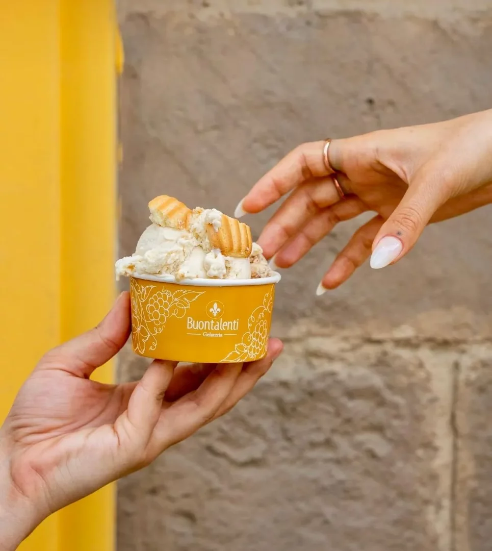 Sarı Buontalenti Gelateria kabında, üzerinde gofret veya kurabiye parçacıkları bulunan ve krem rengi bir gelato dururken, bir el onu tutuyor ve diğer el uzun beyaz tırnaklarıyla nazikçe kaba doğru uzanıyor.