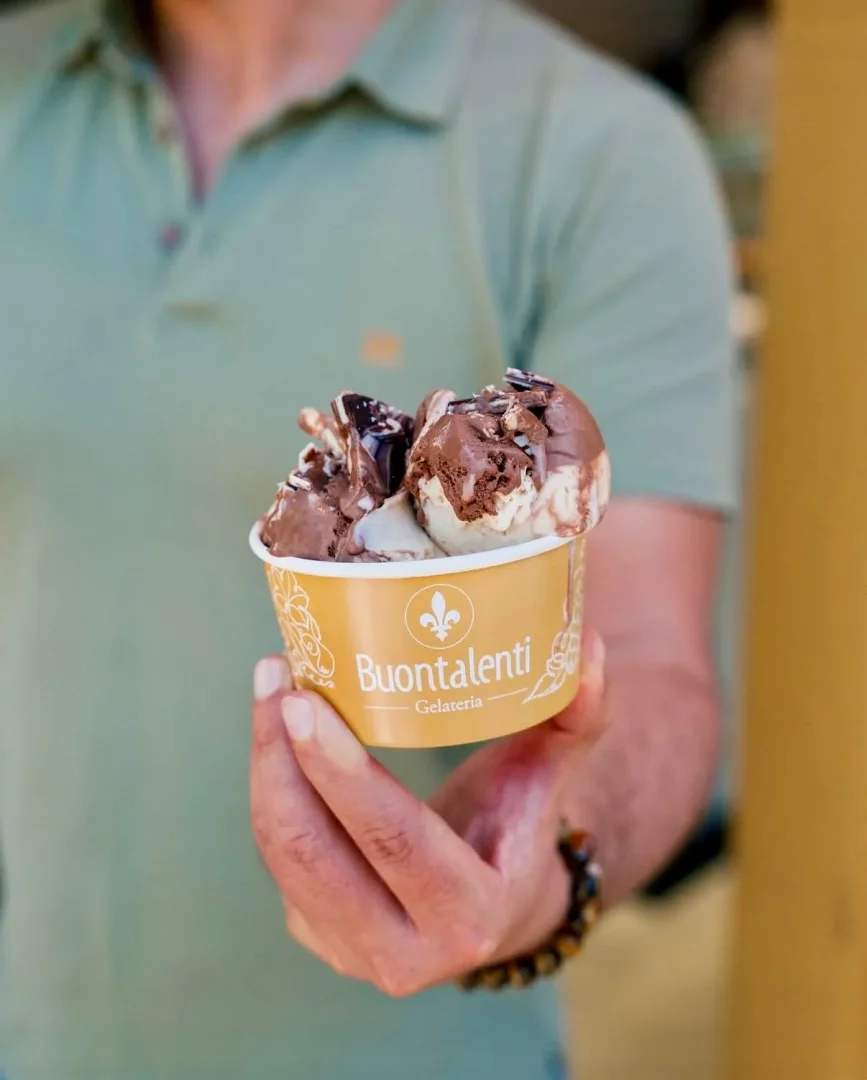 Açık yeşil polo tişörtlü bir kişinin boncuklu bilezik takan eli, üzerinde Buontalenti Gelateria logosu bulunan altın renkli bir kap içinde, çikolata sosu ve parçacıklarıyla süslenmiş, çikolatalı ve vanilyalı dondurmadan oluşan iştah açıcı bir kupayı kameraya doğru uzatıyor.