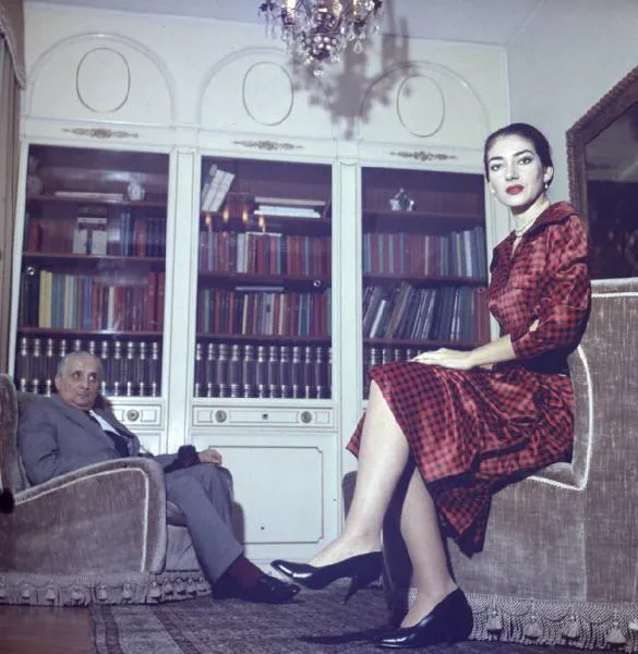 Kırmızı ekose elbisesi, inci kolyesi ve bakımlı saçıyla koltuğun kenarına zarifçe oturmuş Maria Callas'ın, gri takım elbiseli eşi Giovanni Battista Meneghini'nin ona baktığı, arkalarında kitaplarla dolu şık bir kütüphane rafı ve ışıklı bir avizenin bulunduğu 1957 tarihli renkli bir fotoğraf.