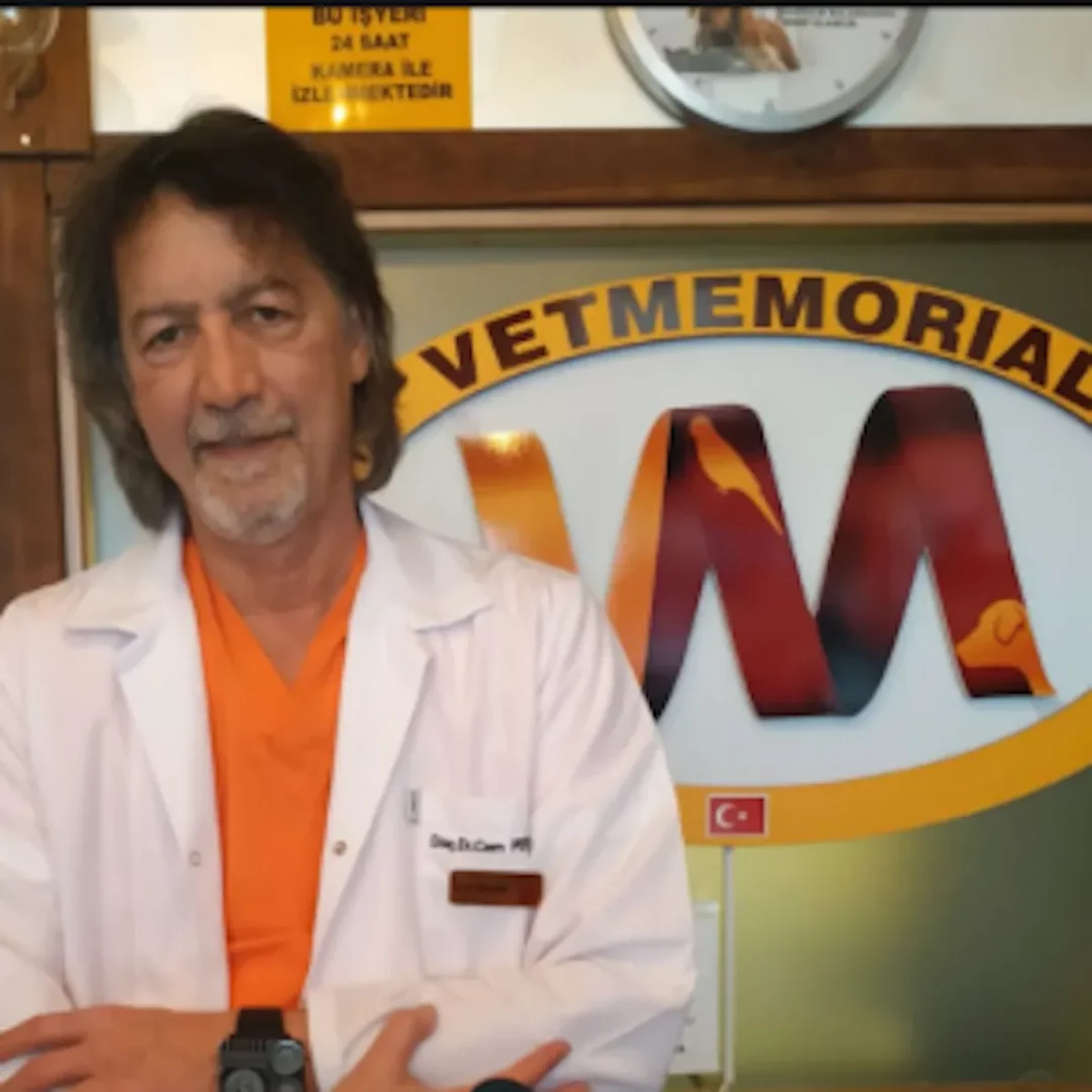 Doç. Dr. Cem Perk, üzerinde beyaz önlüğü ve turuncu içliğiyle kameraya gülümserken, arkasında kuş ve köpek görselleriyle zenginleştirilmiş Vetmemorial kliniği logosu, küçük bir Türk bayrağı ve işyerinin 24 saat kamera ile izlendiğini belirten bir yazı görünen veteriner ortamında duruyor.
