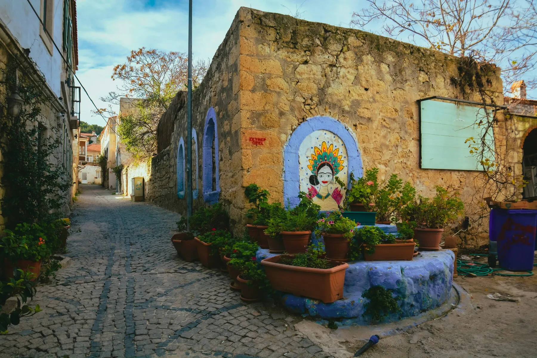 İzmir Çeşme'deki Arnavut kaldırımlı Frida Kahlo Sokağı'nda, sağdaki tarihi taş duvarda canlı renklerle çizilmiş bir Frida Kahlo portresi, mor-mavi boyalı kemerli nişler ve saksılı yemyeşil bitkilerle bezenerek sıcak bir Akdeniz atmosferi sunuyor.