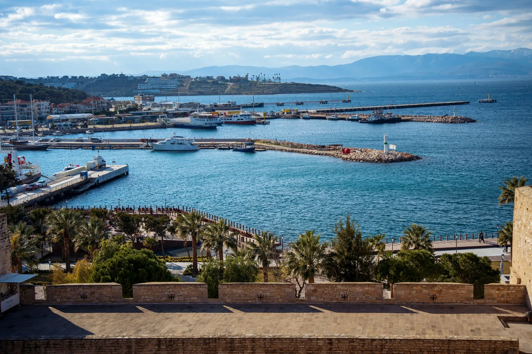 Çeşme, İzmir'in tarihi bir yapıdan çekilmiş kuşbakışı görüntüsünde, masmavi denizde yüzen teknelerle dolu canlı marinası, uzun iskeleleri, palmiye ağaçlarıyla süslenmiş sahil şeridi ve uzaklardaki dağlar net bir şekilde görülüyor.