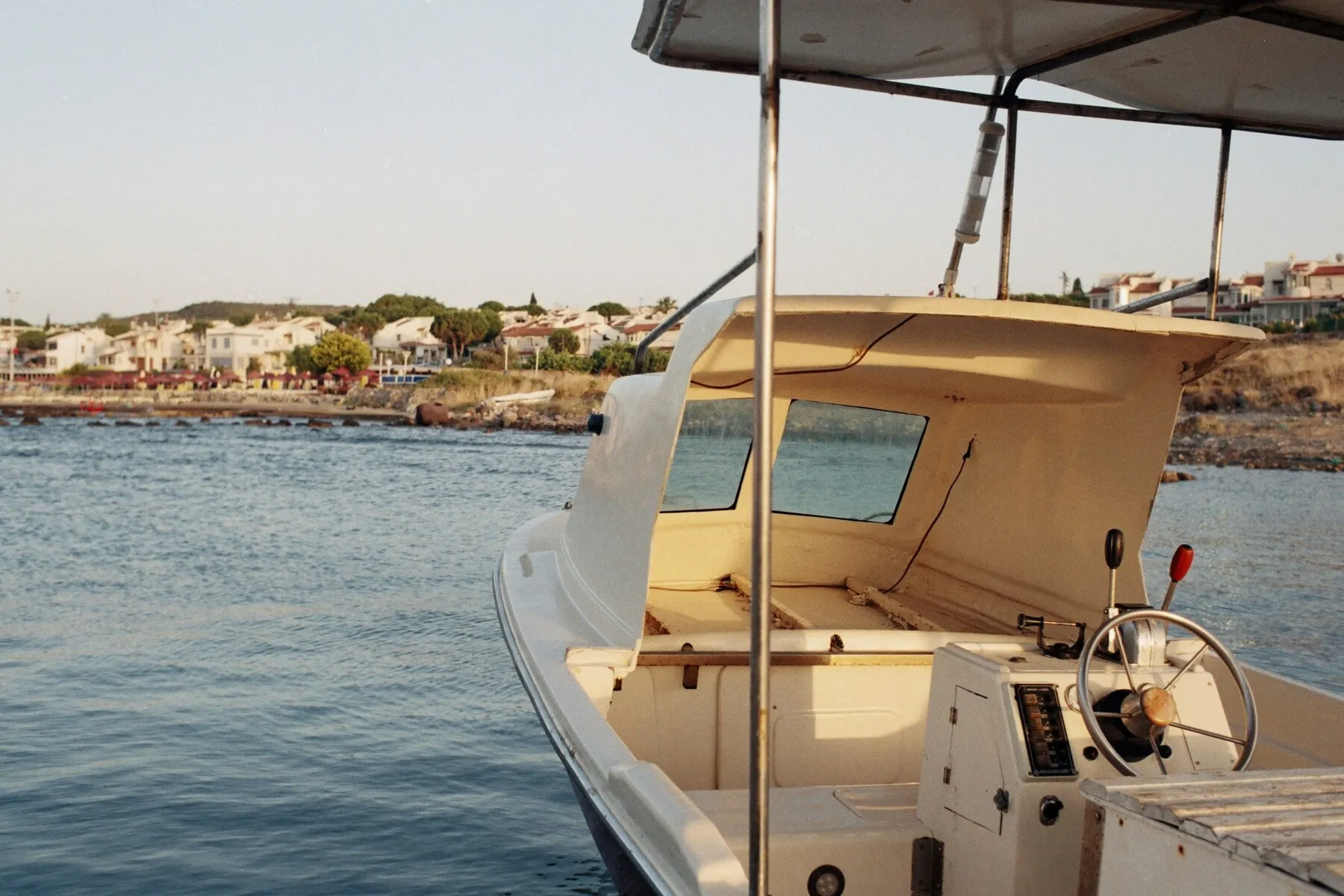 Direksiyonu ve üzeri tenteli, açık renkli bir teknenin güvertesinden sakin mavi sulara bakarken, uzakta Çeşme, İzmir'in sahilindeki beyaz yapılar ve yeşil ağaçlarla dolu yamacın huzurlu manzarası görülüyor.