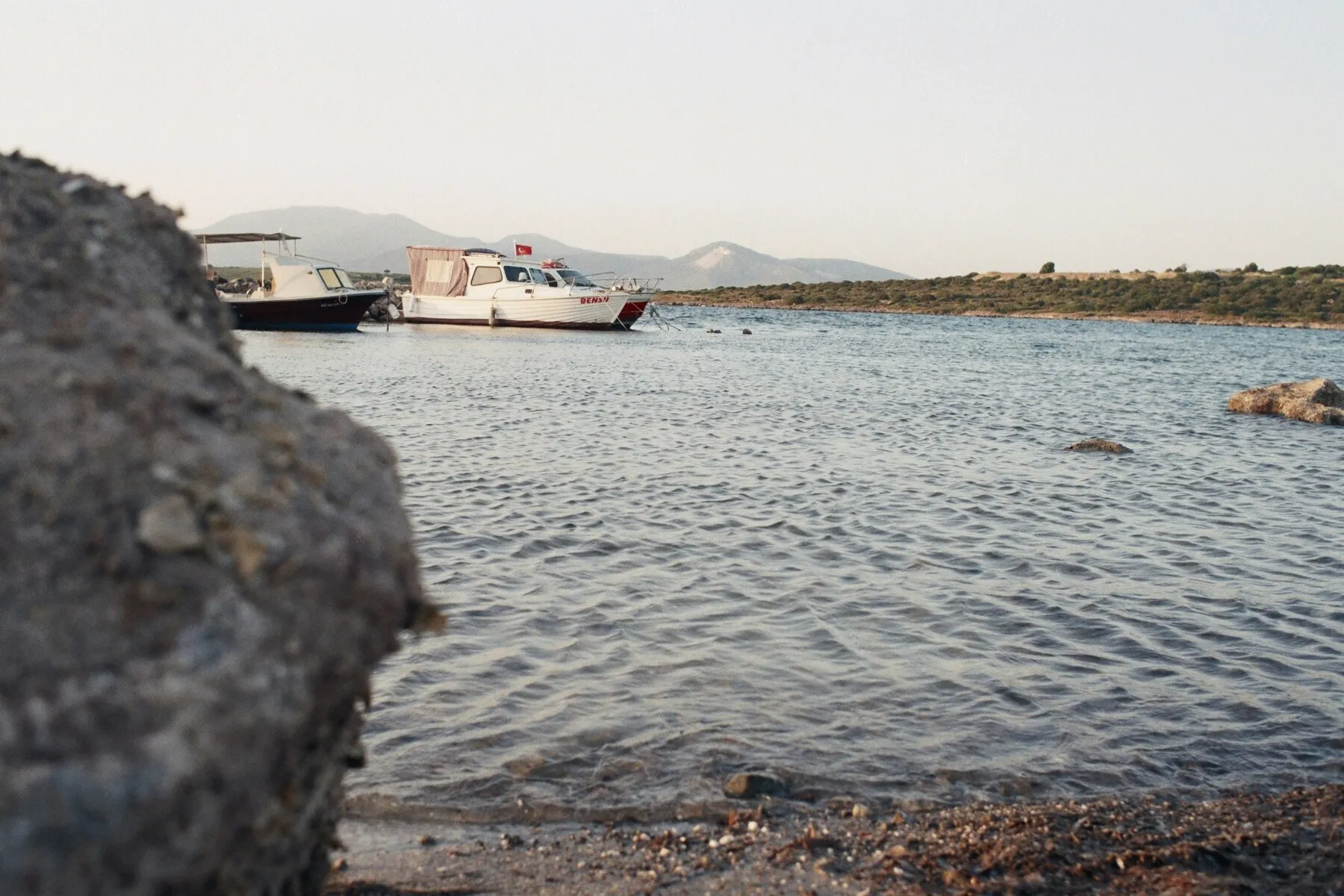 Çeşme ve İzmir'in Ege kıyılarında, sakin denizde demirlemiş, Türk bayraklı beyaz bir tekne ile diğer teknelerin görüldüğü, ön planda bulanık bir kaya ve kumlu kıyının yer aldığı, arkada yeşil tepelerin uzandığı huzurlu bir deniz manzarası.