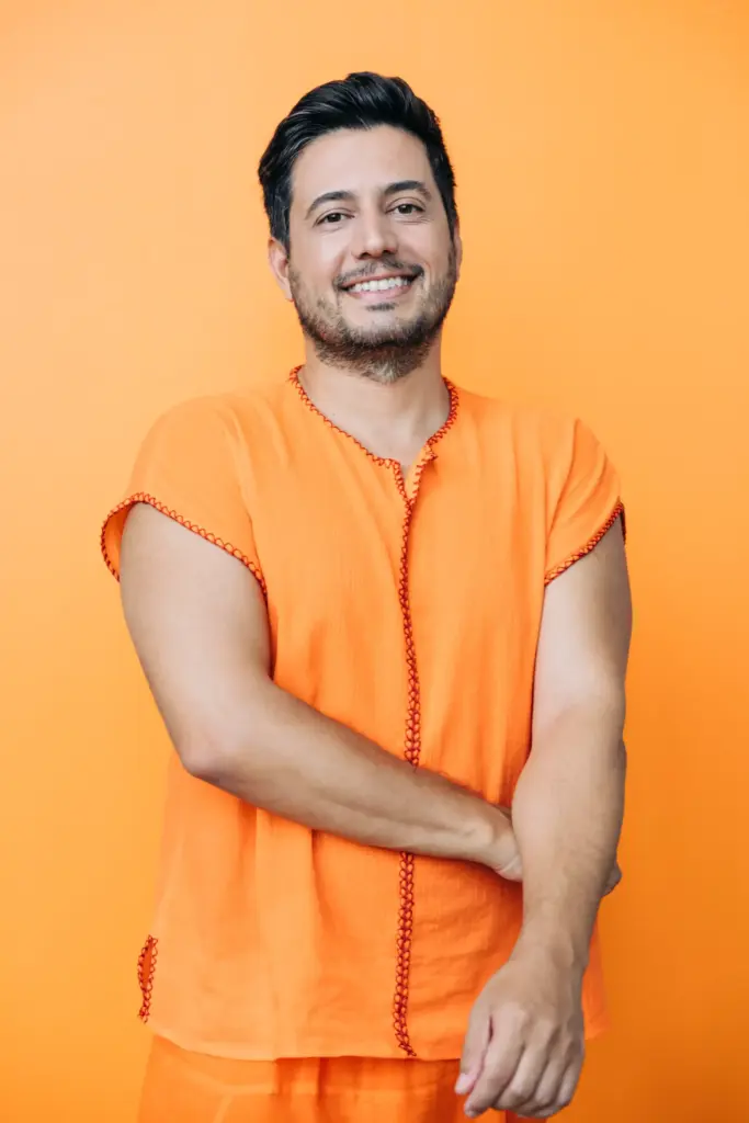 Turuncu arka plan önünde turuncu kıyafet giymiş, gülümseyerek poz veren Çetin Çetintaş, yogi.