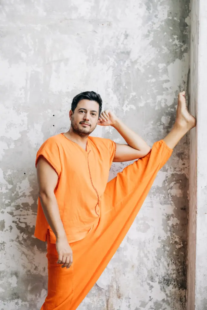 Turuncu kıyafetli Çetin Çetintaş, bir bacağını duvara yaslamış şekilde esneklik gösteren yoga pozu veriyor.