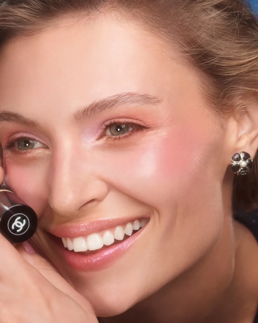 Chanel'in yaz çağıran makyaj görünümünü sergileyen, pembe yanakları, ışıltılı pembe dudakları ve farıyla geniş bir şekilde gülümseyen model, elinde markanın ikonik logosunu taşıyan siyah bir ruj tutarken, kulağındaki inci detaylı çiçek küpesiyle taze ve enerjik bir ifadeye sahip.