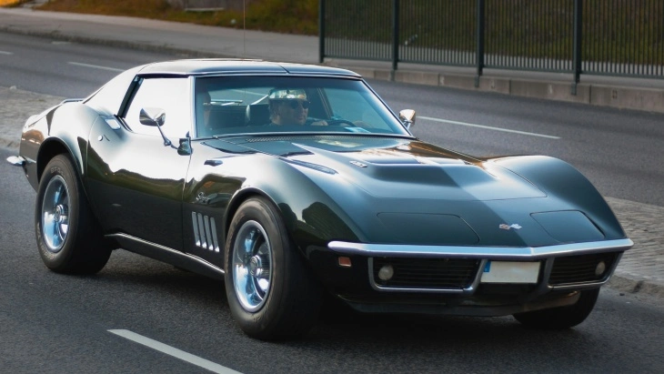 Siyah Chevrolet Corvette Stingray C2, yolda ilerlerken önden yan profilden görüntüleniyor. Kaslı gövde hatları ve ikonik Amerikan spor otomobil tasarımı dikkat çekiyor.