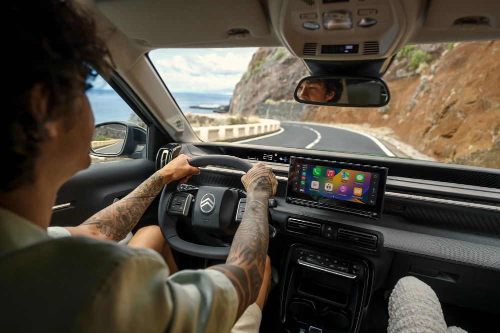 Bir sürücünün kolları dövmeli elleriyle direksiyonu tuttuğu, Citroen e-C3 Aircross elektrikli SUV'un modern ve ferah iç mekan tasarımı, geniş dokunmatik bilgi-eğlence ekranı ve virajlı sahil yolunda ilerlerken görünen denizi gözler önüne seriyor.