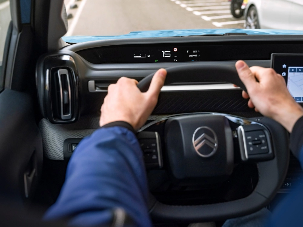 Sürücünün direksiyon başında elleriyle Citroën e-C3 elektrikli SUV'nin modern iç mekanını gösteren POV fotoğrafında, dijital gösterge panelinde 15 km/s hız ve 50 km/s hız sınırı okunurken, sağda navigasyon ekranının bir kısmı görülmektedir.