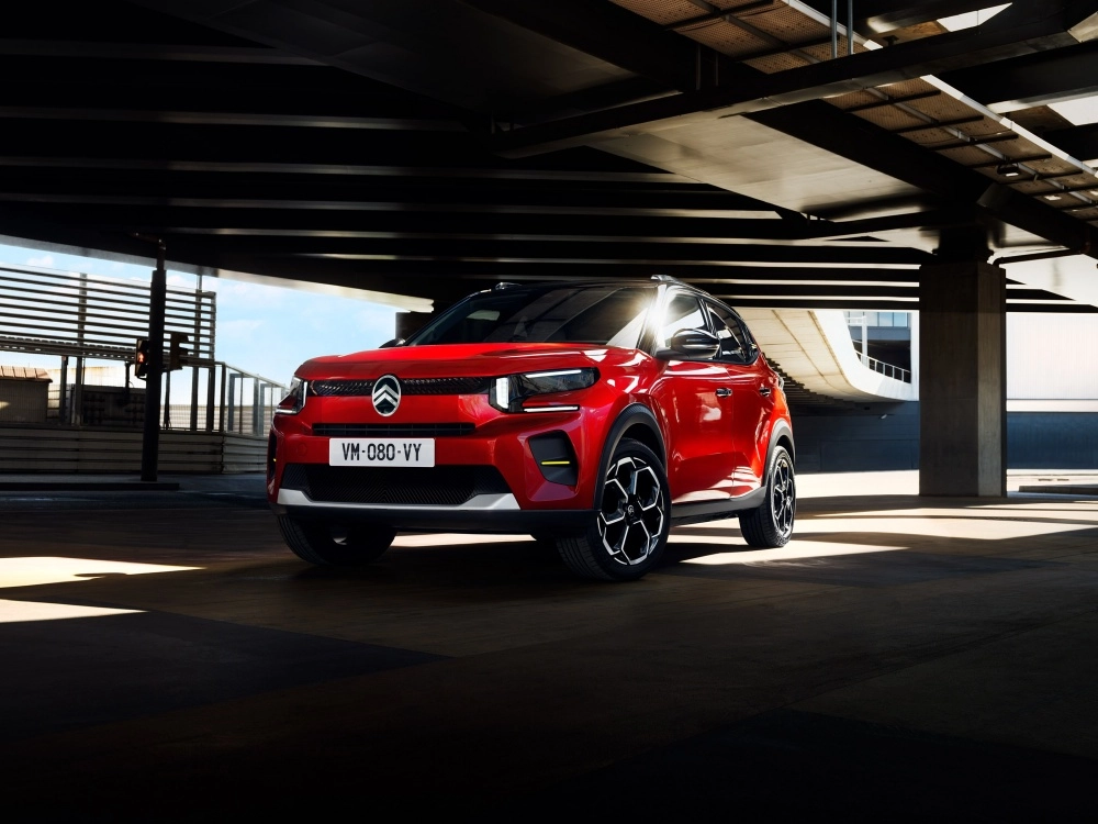 Kırmızı renkteki yeni Citroen e-C3 elektrikli SUV, bir üst geçidin altında, güneşin vurduğu dinamik hatlarıyla dikkat çekici dış mekan tasarımını sergiliyor ve en ucuz elektrikli arabalar arasındaki konumunu görsel olarak yansıtıyor.