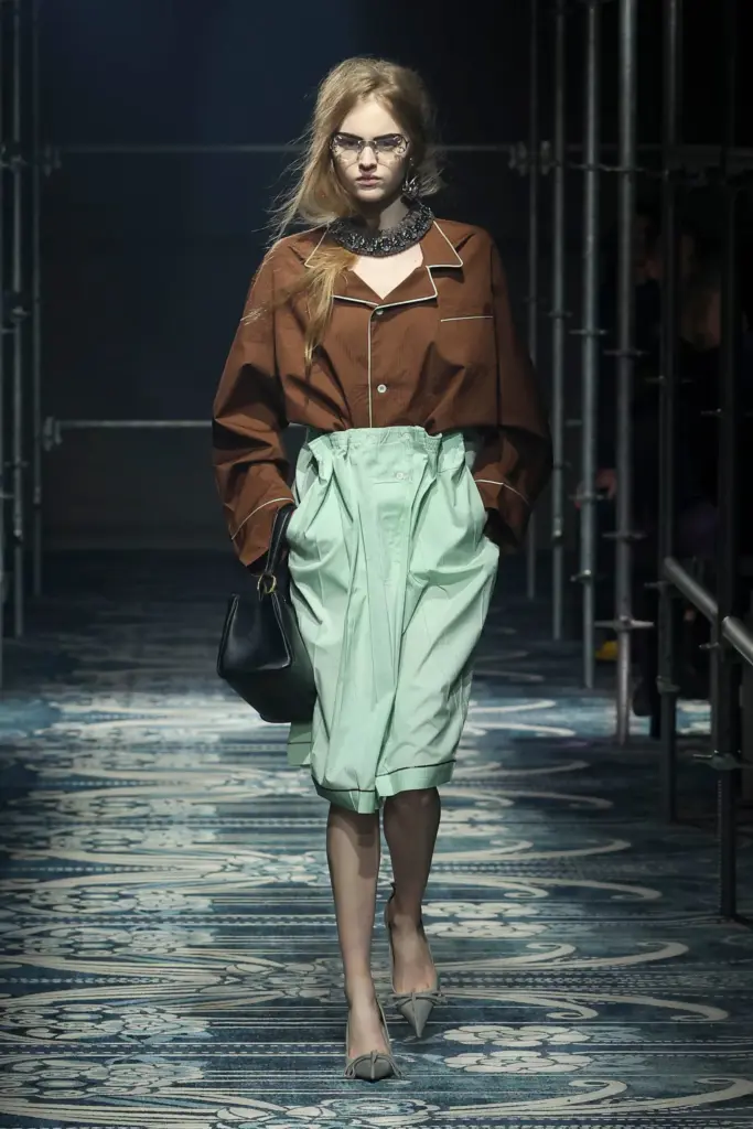 Kahverengi oversized gömlek ve pileli mint yeşili midi etekle yürüyen model, maskülen üstlerle romantik alt parçaların kontrastını yansıtıyor.
