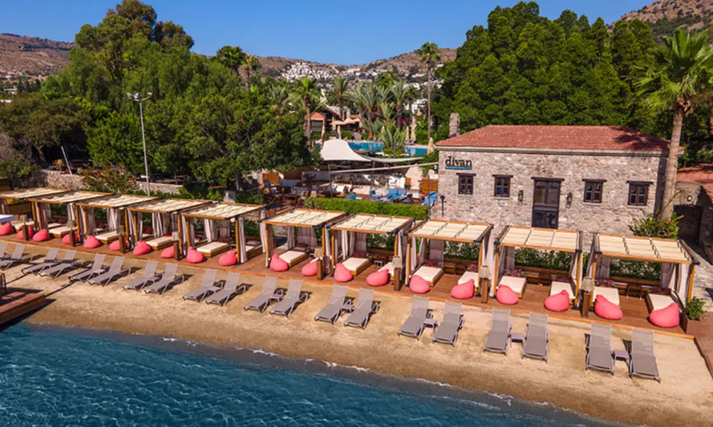Güneşli bir günde, Divan Bodrum'un turkuaz sularına kıyısı olan kumlu sahilinde, bej şezlonglar ile ahşap platform üzerindeki beyaz yataklı ve pembe minderli lüks çardaklar sıralanmış; arka planda Divan yazılı taş bir otel binası ve yemyeşil ağaçlar uzanıyor.