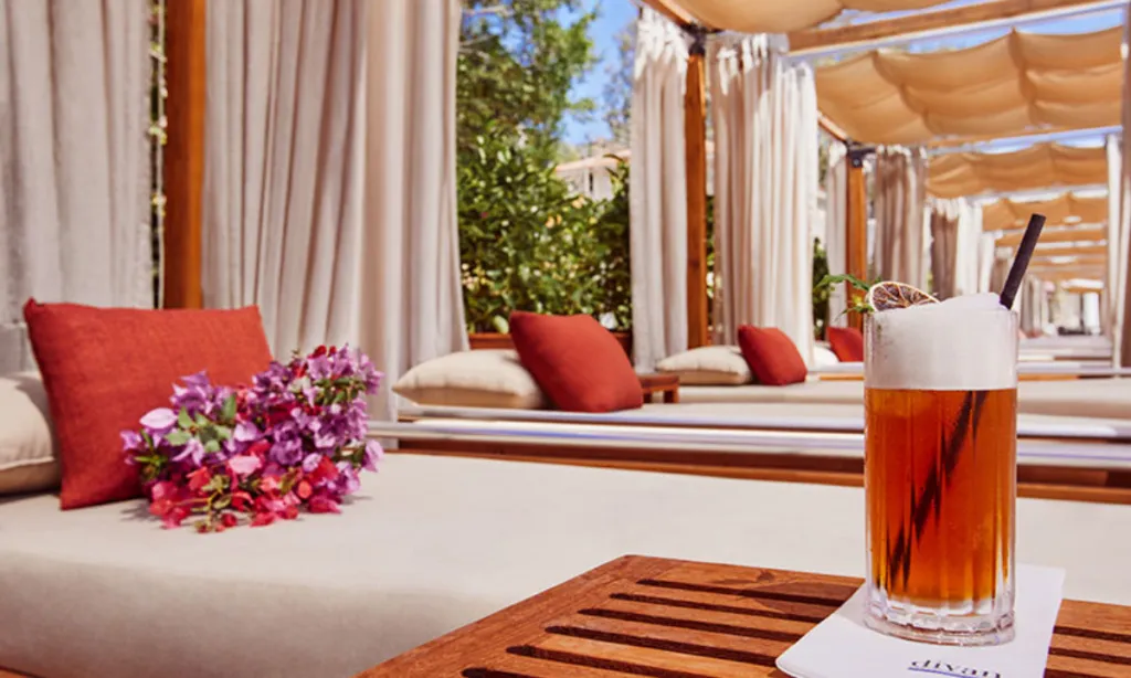 Divan Bodrum Beach'teki gölgelikli lüks dinlenme alanında, ahşap masada narenciye dilimli, köpüklü amber renkli bir kokteyl dururken, mor ve pembe çiçeklerle süslü rahat şezlonglar dingin bir atmosfer yaratıyor.