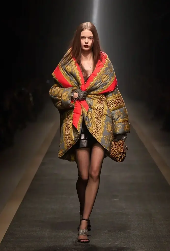 Kırmızı ve altın desenli patchwork puffer ceket, mini deri etek ve koyu makyajla birlikte dramatik maksimalist bir runway görünümü sunuyor.
