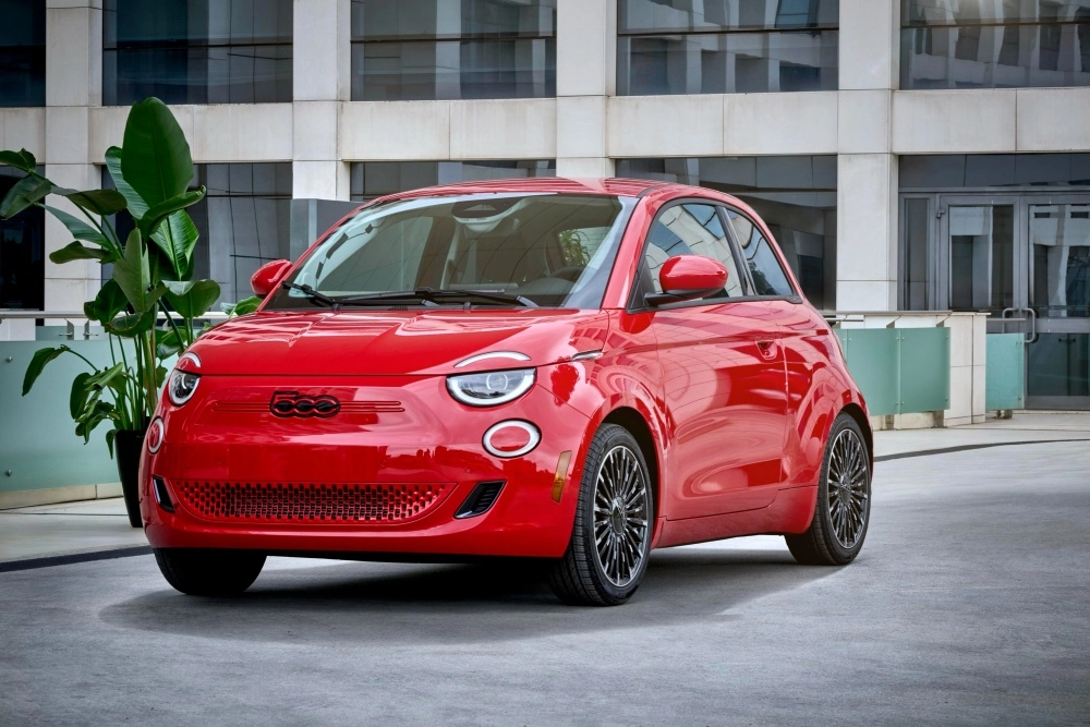 Kırmızı renkli bir Fiat 500e elektrikli şehir içi hatchback otomobil, modern bir cam cepheli binanın önünde, solunda büyük yeşil bir bitkiyle dış mekanda hafif çapraz açıyla park edilmiş, belirgin yuvarlak farları ve çok kollu koyu renk jantlarıyla dikkat çeken şık dış mekan tasarımını gözler önüne seriyor.