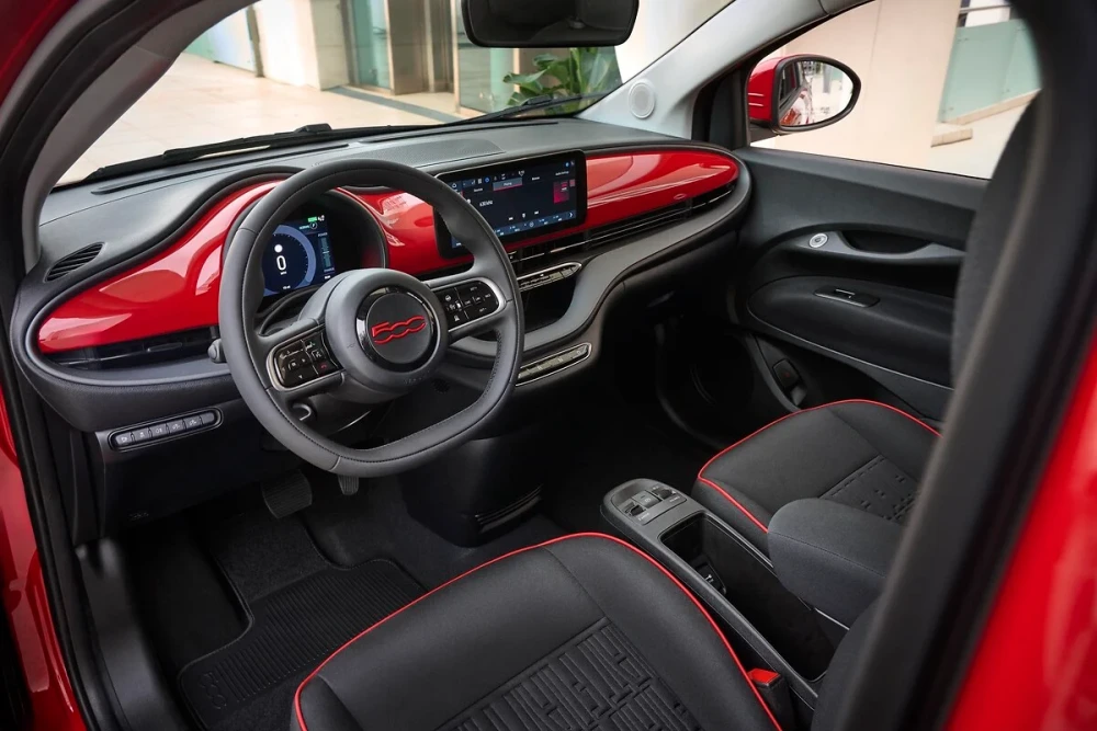 Kırmızı renk detaylarıyla dikkat çeken Fiat 500e'nin modern iç mekan tasarımı, sürücü koltuğundan görünen siyah direksiyon simidi, dijital gösterge paneli ve merkezi bilgi-eğlence ekranıyla şık ve ergonomik bir görünüm sunuyor.