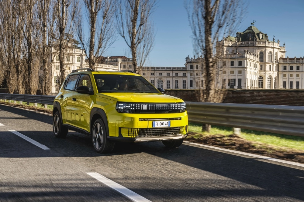 Canlı sarı rengiyle dikkat çeken, geleceğe dönük tasarımlı LED farlara ve modern bir ön ızgaraya sahip, ilk elektrikli otomobil modellerinden biri olan Fiat Grande Panda SUV, tarihi bir yapının ve ağaçların önünden geçerek yol üzerinde ilerlerken görünüyor.