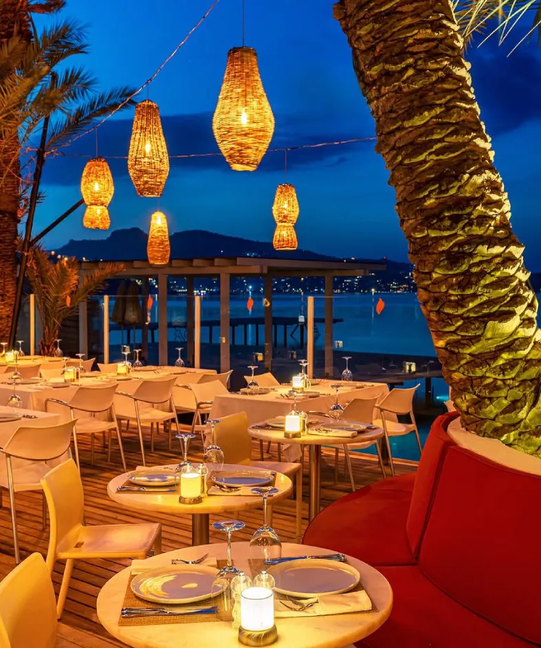 Bodrum'daki Flamm Beach restoranının akşam yemeği atmosferini yansıtan bu görüntüde, ahşap zemine yayılmış, hasır lambalarla sıcak bir şekilde aydınlatılmış birçok yuvarlak masa ve sandalyeler, arka planda lacivert deniz ve dağ manzarasıyla buluşuyor.