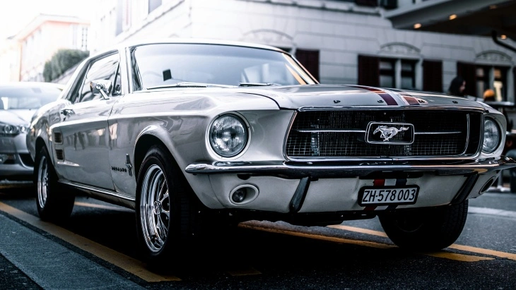 Beyaz renkli klasik Ford Mustang Fastback, şehir sokaklarında park halinde. Ön ızgarasında Mustang logosu ve ikonik muscle car tasarımıyla dikkat çekiyor.