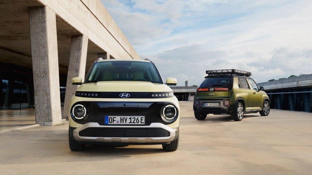 Önde açık sarı renkteki Hyundai Inster kompakt SUV elektrikli otomobilin belirgin ön ızgarası ve yuvarlak farları görünürken, arkada yeşil renkli aracın piksel desenli stop lambaları ve portbagajlı modern dış tasarımı mimari bir alanda sergileniyor.