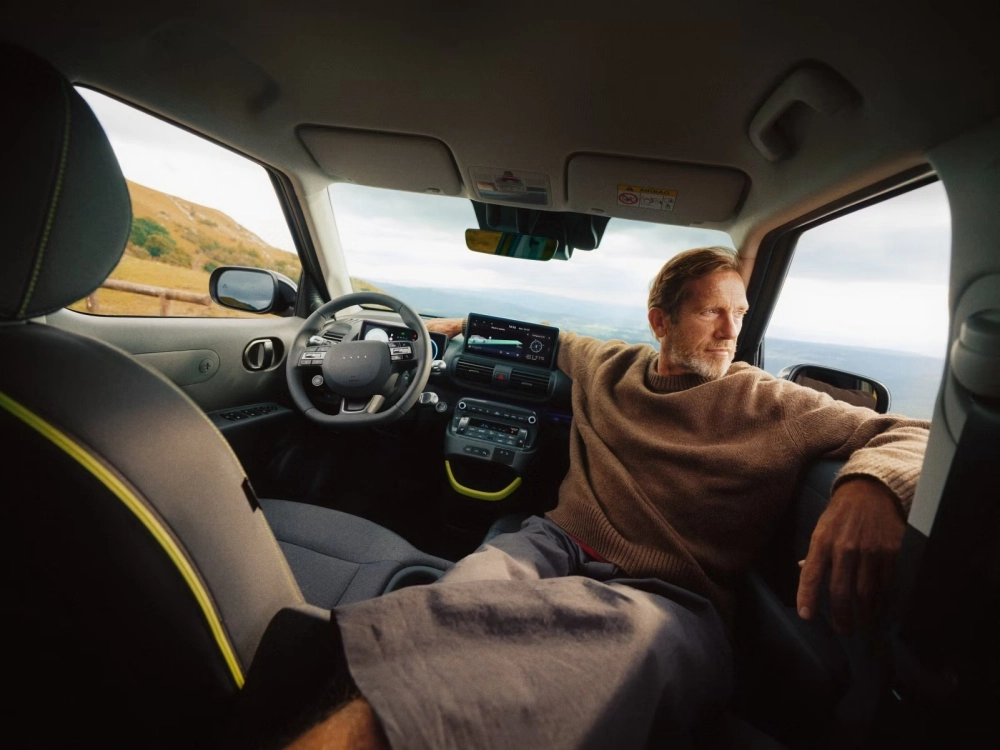 Hyundai Inster elektrikli kompakt SUV'un modern iç mekan tasarımı, kahverengi kazaklı bir adamın yolcu koltuğunda pencereden dışarıdaki doğal manzarayı seyretmesiyle rahat ve keyifli bir yolculuk deneyimini sergiliyor.