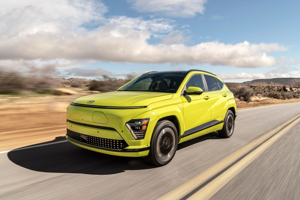 Sarı renkli yeni kasa Hyundai Kona Electric elektrikli SUV, çöl benzeri kuru bir arazideki asfalt yolda hareket halinde, aracın çarpıcı dış mekan tasarımı ve canlı rengi gökyüzündeki bulutların altında öne çıkıyor.