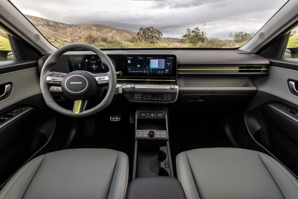 Geniş ön camdan dağlık bir arazinin göründüğü, Hyundai Kona Electric modelinin modern iç mekan tasarımını gösteren bu fotoğrafta, gri delikli koltuklar, sarımsı yeşil vurgulara sahip çift ekranlı dijital kokpit, şık direksiyon simidi ve kullanışlı orta konsol detaylarıyla elektrikli SUV'un ferah ve teknolojik atmosferi vurgulanıyor.