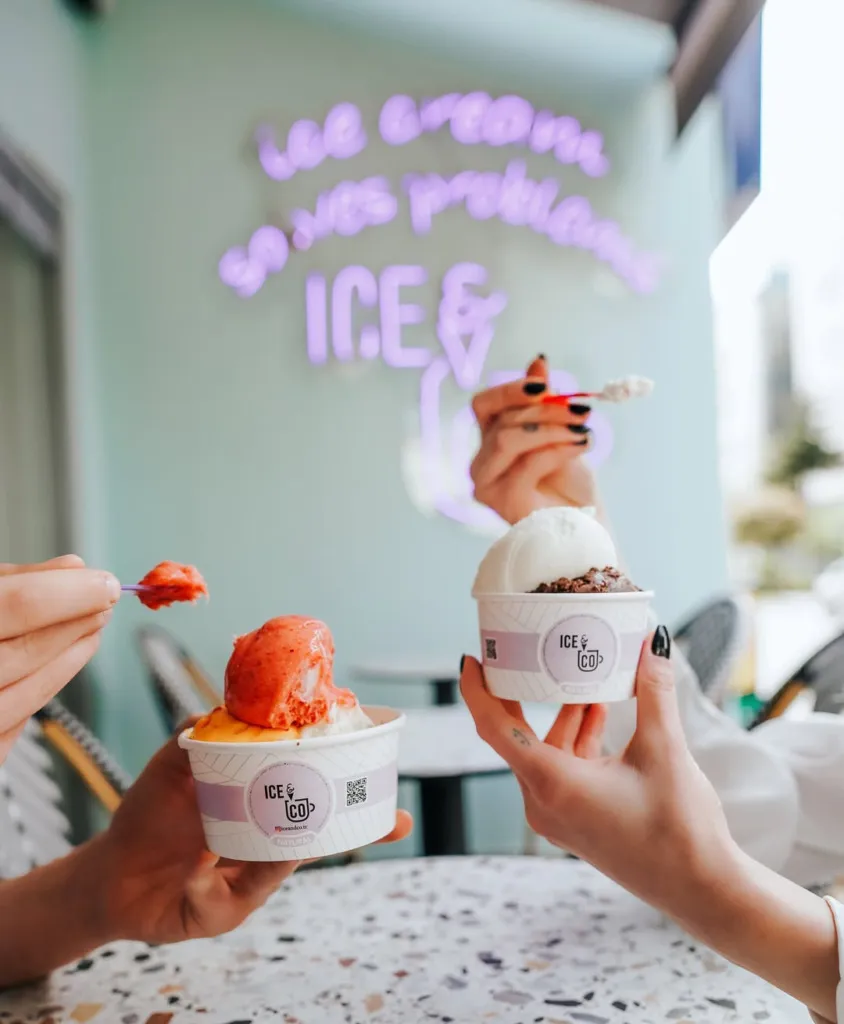 ICE & CO dondurma dükkanında iki el, çilekli ve portakallı bir dondurma ile çikolatalı ve vanilyalı başka bir dondurma kabı tutarken, üçüncü bir el kaşıklı dondurma servis ediyor; arka planda ise ICE & CO'nun neon tabelası dikkat çekiyor.