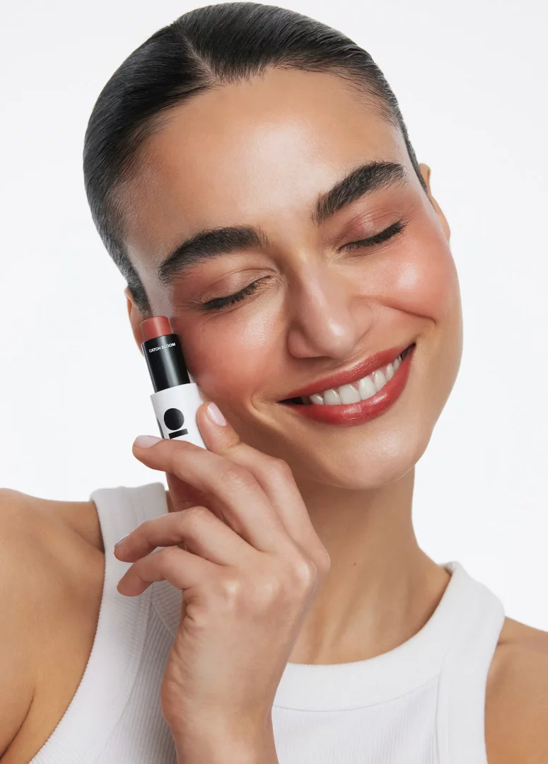 Mutlu ve gözleri kapalı gülümseyen bir kadın, beyaz tişörtüyle, yanağının hemen yanında, doğal ve ışıltılı bir görünüm veren ino beauty stick ruj allık ürününü tutuyor, makyajıyla sağlıklı ve canlı bir güzellik sergiliyor.