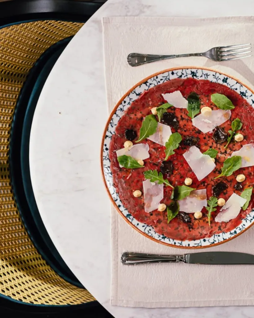 Beyaz mermer bir masanın üzerinde, desenli kenarlı bir tabakta roka yaprakları, ince parmesan dilimleri ve küçük fındıklarla zenginleştirilmiş kırmızı et carpaccio tabağı, zarif çatal ve bıçak takımıyla birlikte şık bir Isola Manzara Garden Bodrum sunumunu yansıtıyor.