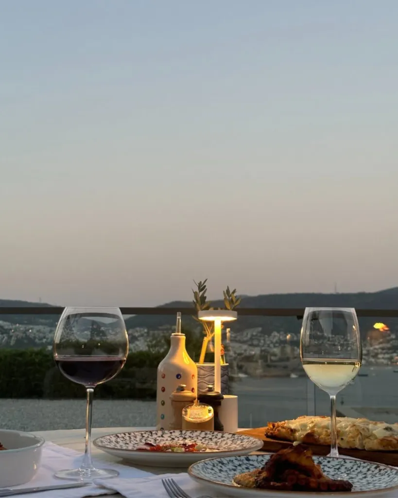 Bodrum seyahat rehberi: Bodrum'da gün batımında şık bir teras restoranında, iki kadeh şarap, zeytinyağı şişesi ve çeşitli lezzetlerle donatılmış masanın önünde, tepelik binalarla çevrili körfez ve şehrin ışıkları eşliğinde keyifli bir akşam yemeği manzarası.