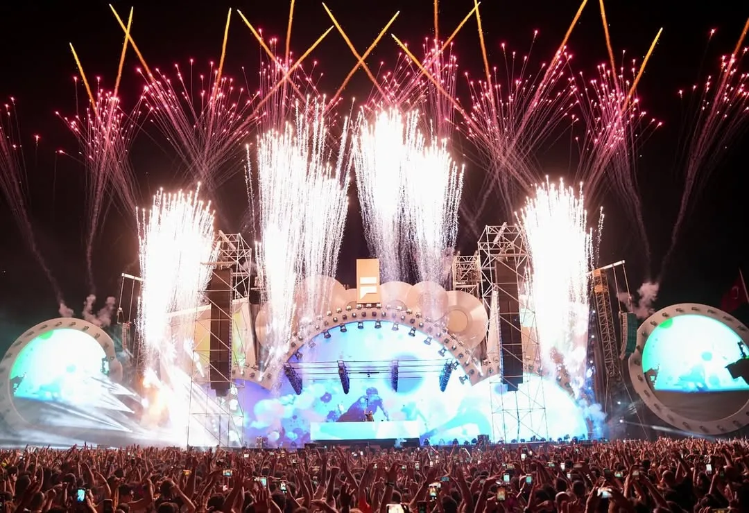 Gecenin karanlığında, İstanbul Festivali'nin Yenikapı'daki büyük konser alanında, havai fişek gösterisiyle aydınlanan sahne önünde toplanmış coşkulu binlerce izleyici ellerini havaya kaldırarak bu muhteşem açık hava etkinliğinin tadını çıkarıyor.