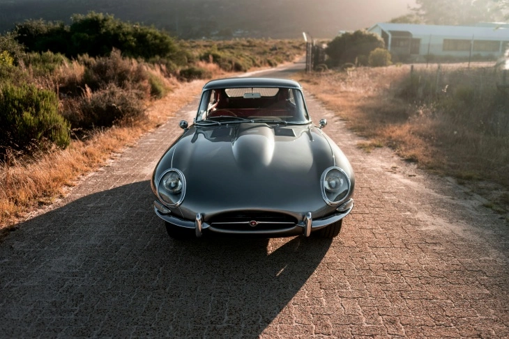 Gümüş renkli Jaguar E-Type, kırsal bir yolda önden görünümüyle sergileniyor. Uzun motor kaputu ve zarif tasarımıyla dünyanın en güzel otomobillerinden biri kabul edilen klasik model.