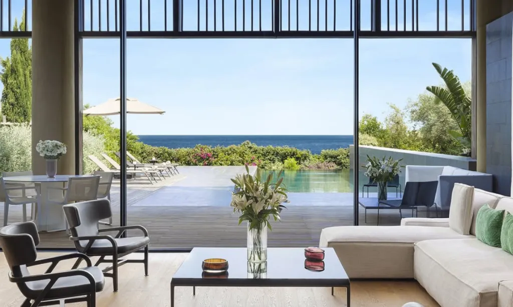 Modern ve aydınlık bir oturma odasından denizin, havuzun ve yemyeşil Akdeniz bitki örtüsünün eşsiz panoramik manzarasını sunan lüks bir otel süiti veya villası, Bodrum'daki konforlu bir tatil deneyimini vurguluyor.