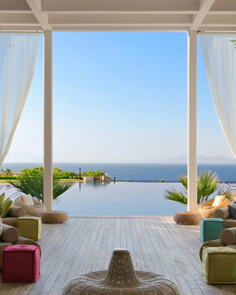 Kempinski Bodrum'un açık hava terasından görünen, şık ahşap zemini, renkli minderli oturma gruplarını ve palmiye ağaçlarıyla çevrili sonsuzluk havuzunun ötesinde uzanan masmavi Ege Denizi ile berrak gökyüzünün birleştiği huzur dolu bir manzara.