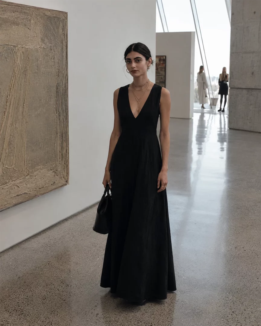 Modern bir sanat galerisi ortamında, şık ve derin V yaka siyah uzun bir elbise giymiş, zarif altın kolye katmanları ve küpelerle süslenmiş, elinde küçük siyah bir çanta tutan genç bir kadın, gün batımı davetleri için sofistike ve etkileyici bir tarz sunuyor.